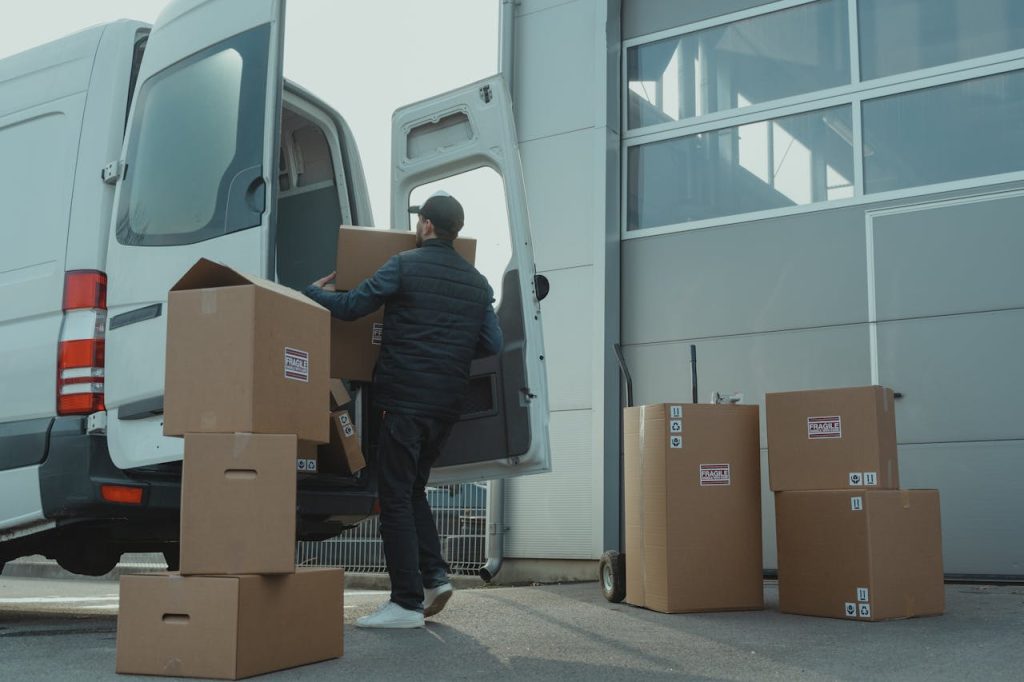 man-in-blue-jacket-and-blue-denim-jeans-sitting-on-brown-cardboard-box-6169056 A delivery man unloading cardboard boxes from a van at a warehouse during the day.
